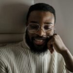 Home A relaxed bearded man with glasses smiles warmly while leaning on his hand indoors.